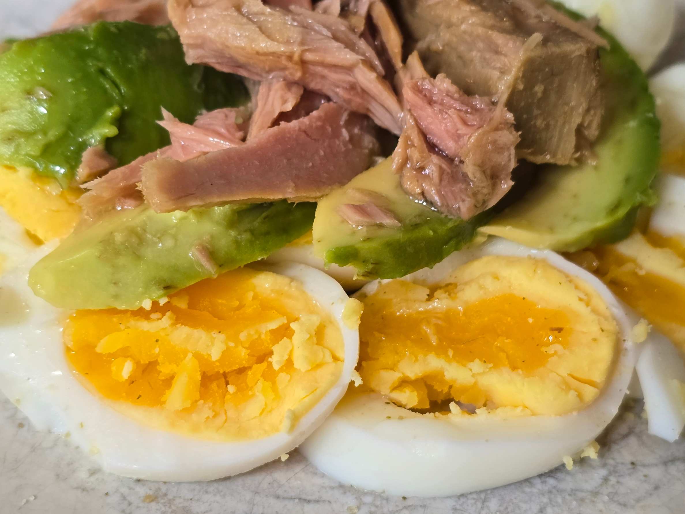 Close-up of slices of hard boiled eggs with slices of avocado on and chunks of tuna on top on a white plate