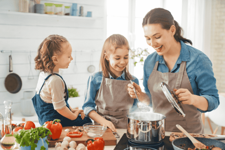 A woman cooking at home with joy surrounded by two daughters smiling
