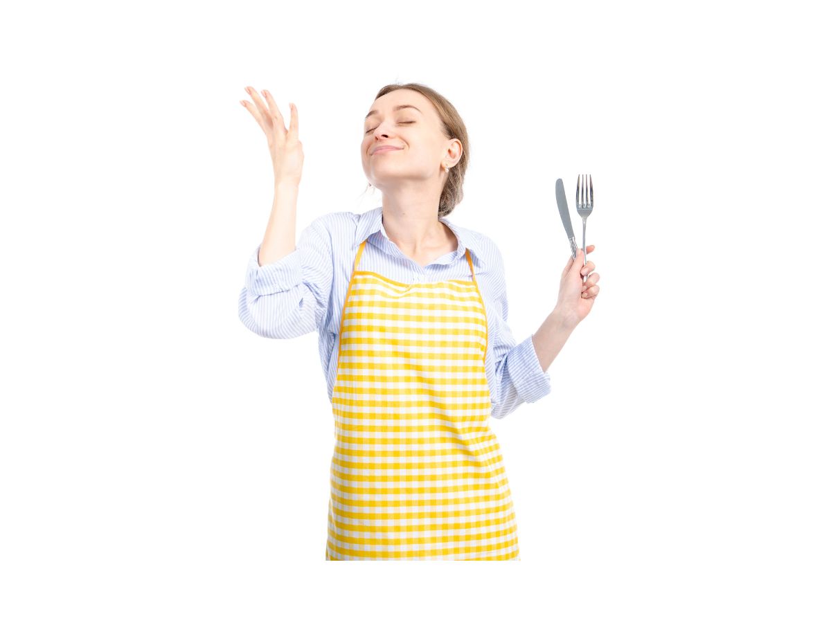 Young woman wearing yellow apron holding a knife and fork in one hand smiling with eyes closed imaging the delicious food she prepared for herself