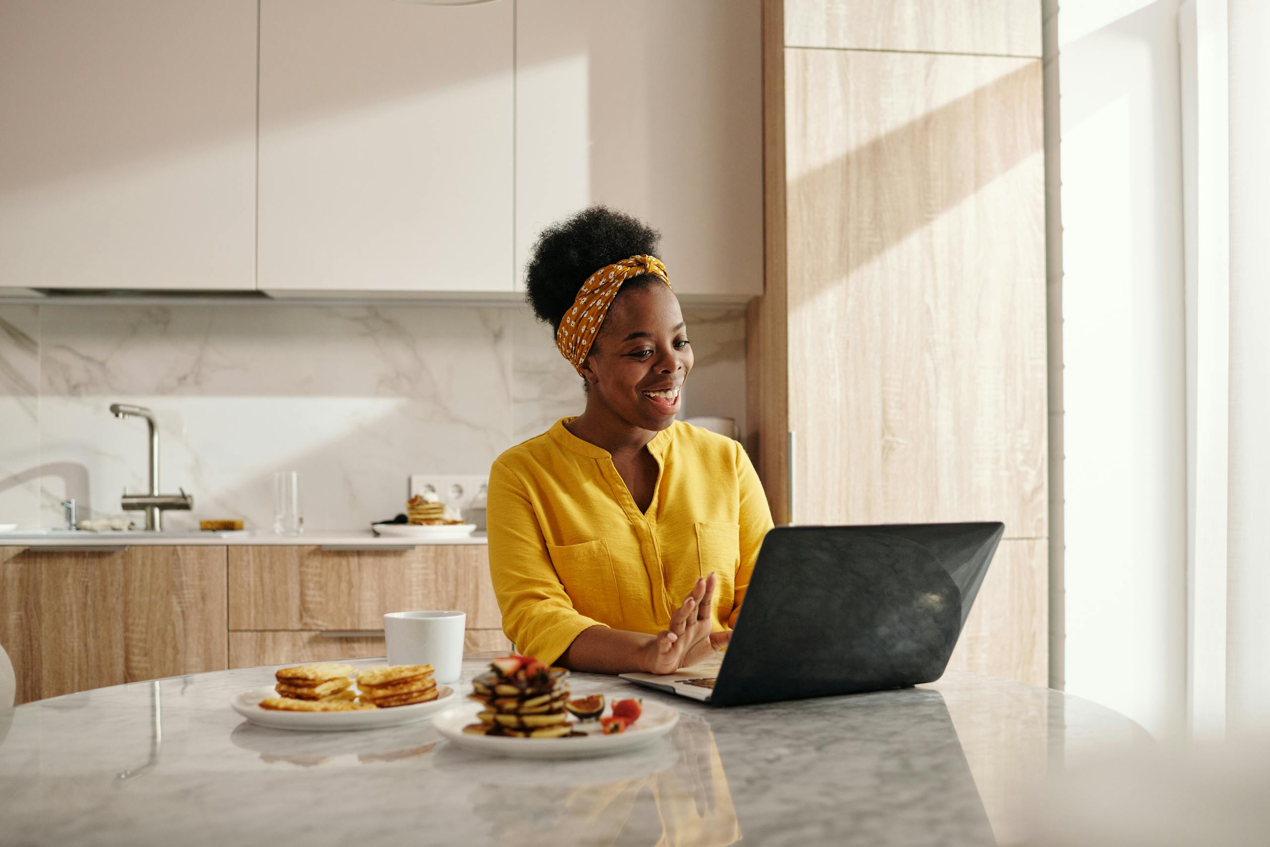 Smiling woman in yellow enjoys breakfast while using a laptop in her sunny kitchen.